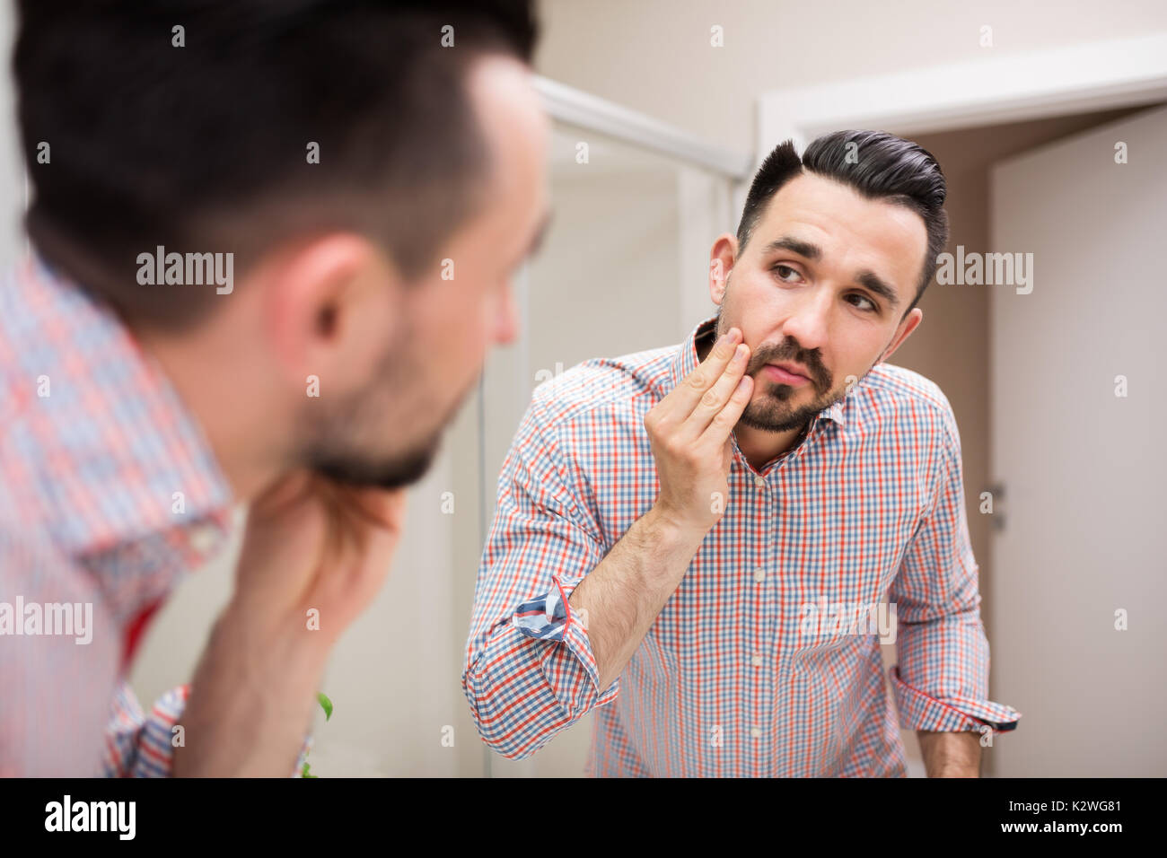 Uomo bello esaminando la sua faccia nello specchio. L uomo è indossare Camicia casual con arrotolato le maniche. Egli è in piedi nel bagno interno. Foto Stock