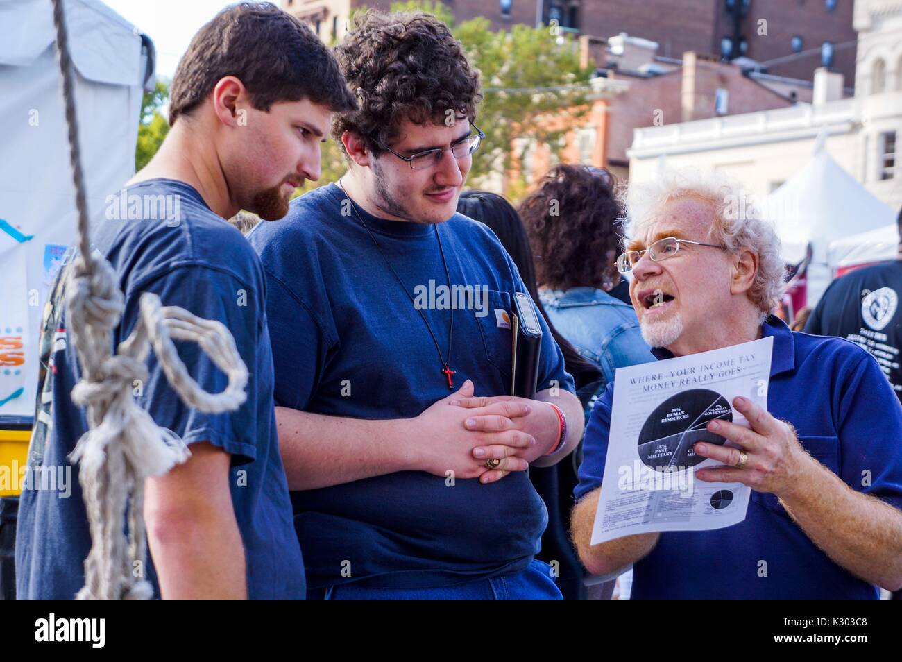 Un uomo anziano con gli occhiali legge da un opuscolo sulle imposte sul reddito di due giovani uomini che indossano croci e azienda bibbie, durante la Baltimore Book Festival, Baltimore, Maryland, Settembre, 2013. La cortesia Eric Chen. Foto Stock