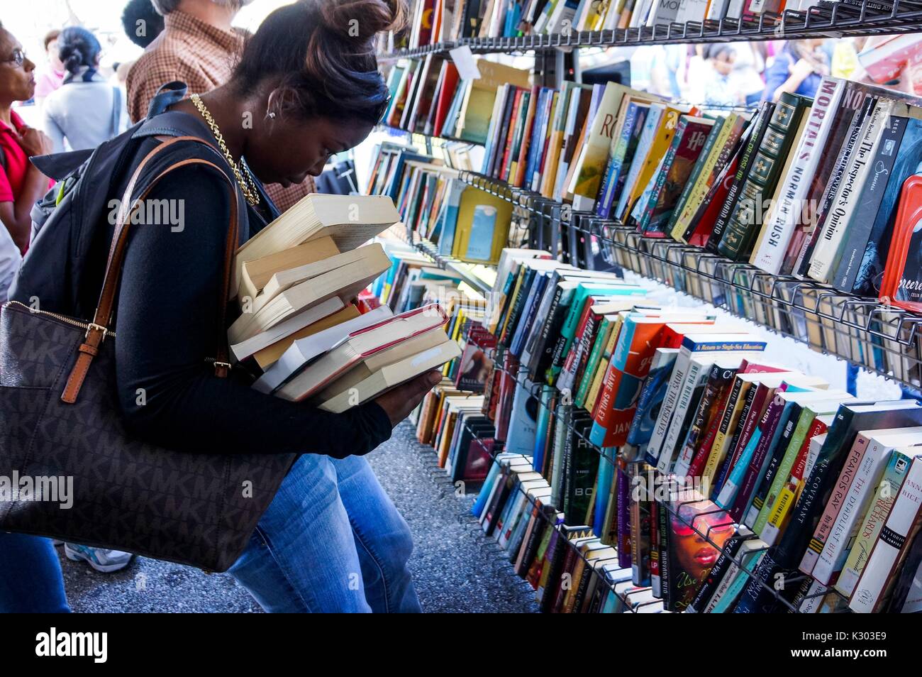Una giovane donna fa fatica a tenere una inclinazione pila di libri nelle sue braccia come lei osserva i titoli di libri usati su un ripiano durante il Baltimore Book Festival, Baltimore, Maryland, 2013. La cortesia Eric Chen. Foto Stock