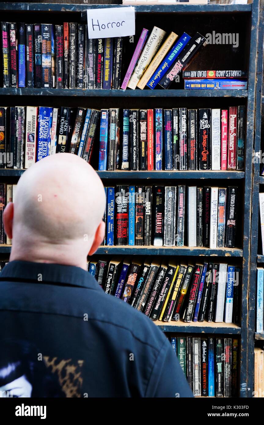 Un uomo con testa calva sembra fino ad un ripiano di usato libri in brossura etichettato 'Horror, ' durante la Baltimore Book Festival, Baltimore, Maryland, Settembre, 2013. La cortesia Eric Chen. Foto Stock