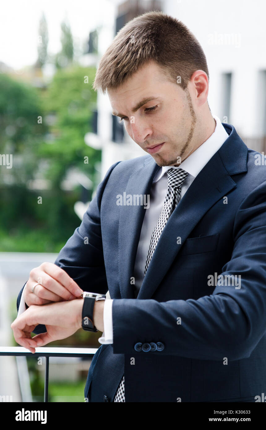 Imprenditore rilassato in piedi sul balcone del suo appartamento e guarda l'orologio Foto Stock