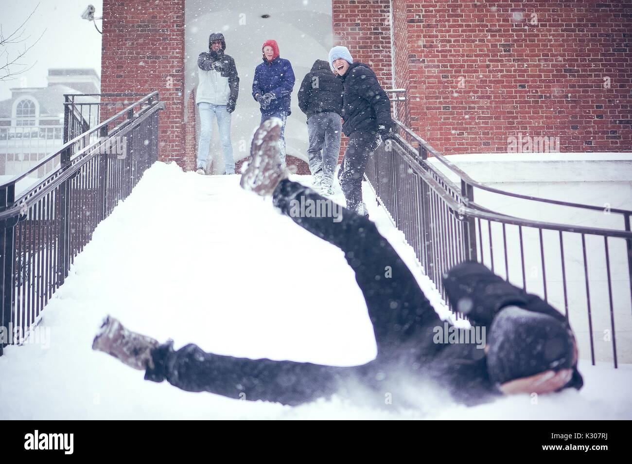 Uno studente scorre verso il basso una scale innevato con le gambe in aria e dei bracci che copre la sua testa, mentre gli altri studenti in marcia sulla neve il sorriso e allegria in cima alle scale, in un giorno di neve alla Johns Hopkins University, Baltimora, Maryland, 2016. Foto Stock