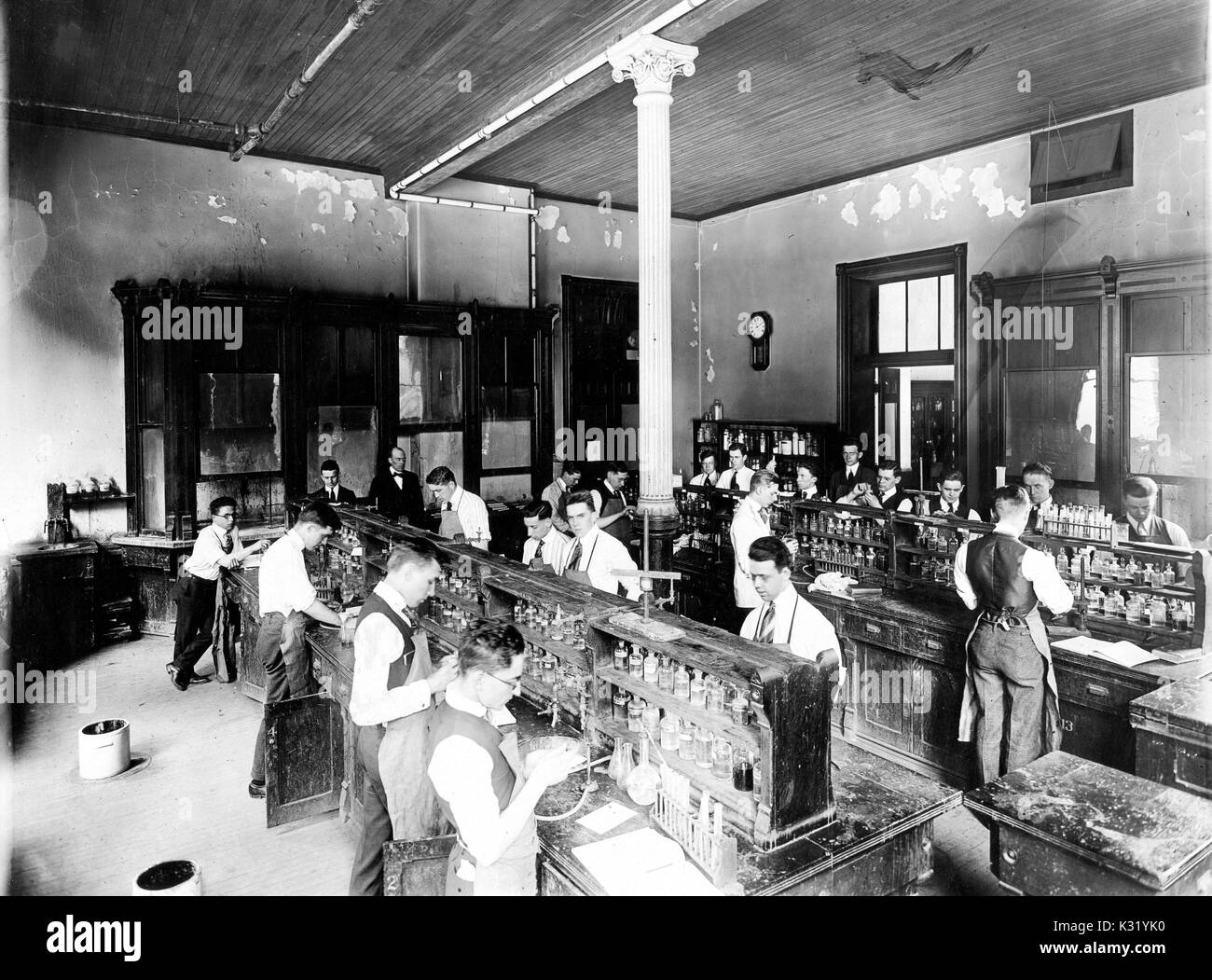 Fotografia in scala di grigi dei maschi di studenti di chimica lavora in un laboratorio di un edificio chimica dal vecchio Campus della Johns Hopkins University, allineati in corrispondenza di panche con bicchieri e provette, indossando i legami al di sotto dei grembiuli, Baltimore, Maryland, 1900. Foto Stock