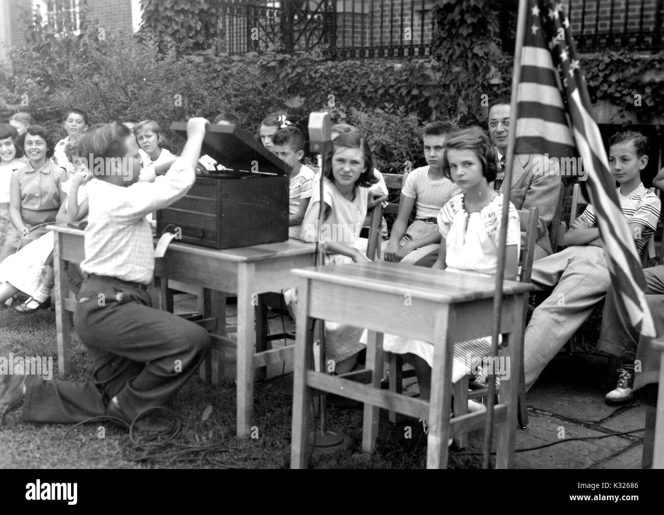 Al giorno di chiusura attività per una dimostrazione elementare Scuola presso la Johns Hopkins University, gli studenti si siedono a sedie in fila mentre un compagno di classe prepara un lettore di musica e una bandiera americana prima per il canto dell'inno nazionale, al di fuori del quadrangolo di fronte un edera-coperta edificio del campus, Baltimore, Maryland, luglio 1950. Foto Stock