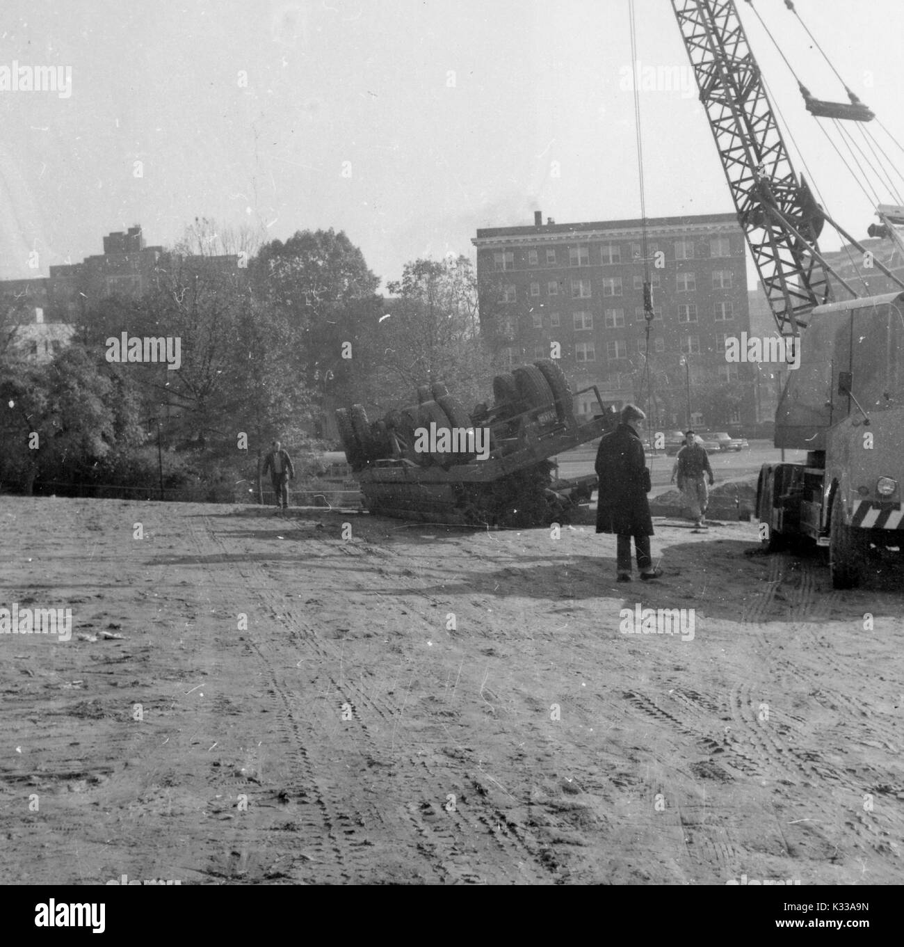 Durante le prime fasi di costruzione del Milton S Eisenhower biblioteca presso la Johns Hopkins University, lavoratori utilizzano una grande gru per tentare di correggere un carrello ribaltata in posizione verticale, con tre lavoratori a piedi attorno al terreno fangoso, davanti a McCoy Hall, un dormitorio underclassman edificio su Charles Street, Baltimore, Maryland, 1963. Foto Stock