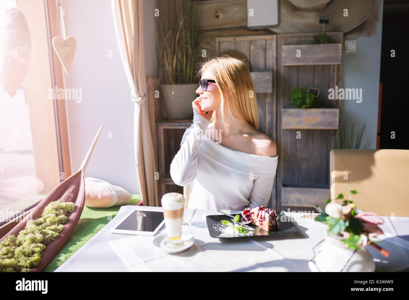 Una foto di una giovane e bella donna di occhiali da sole seduti al tavolo del bar. Sta parlando al cellulare e guardando fuori dalla finestra. Foto Stock