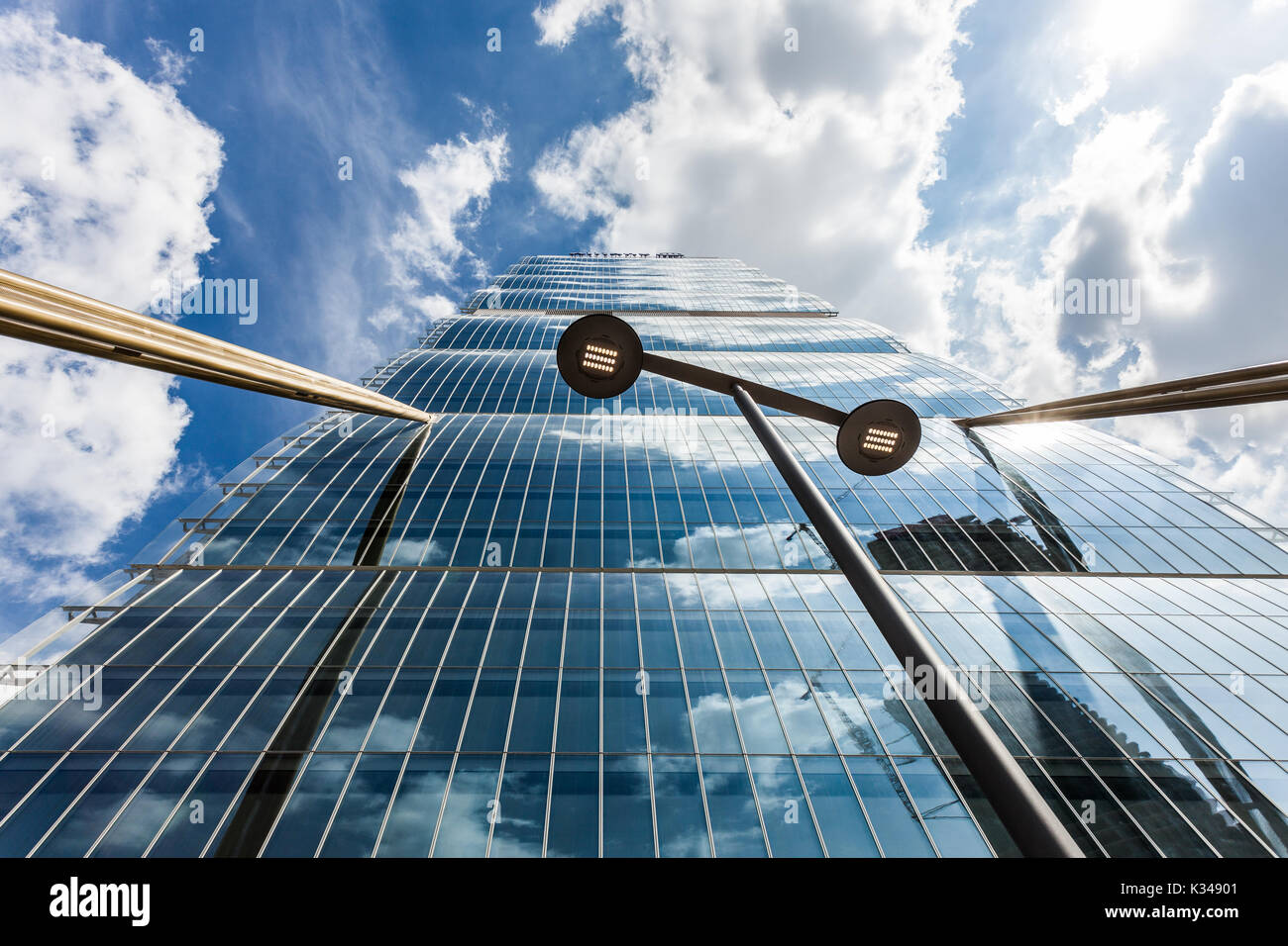 Vista ad angolo basso di un grattacielo di vetro che riflette le nuvole e il cielo, incorniciato da travi dorate e un lampione, simbolo dell'architettura urbana e dell'innovazione. Foto Stock