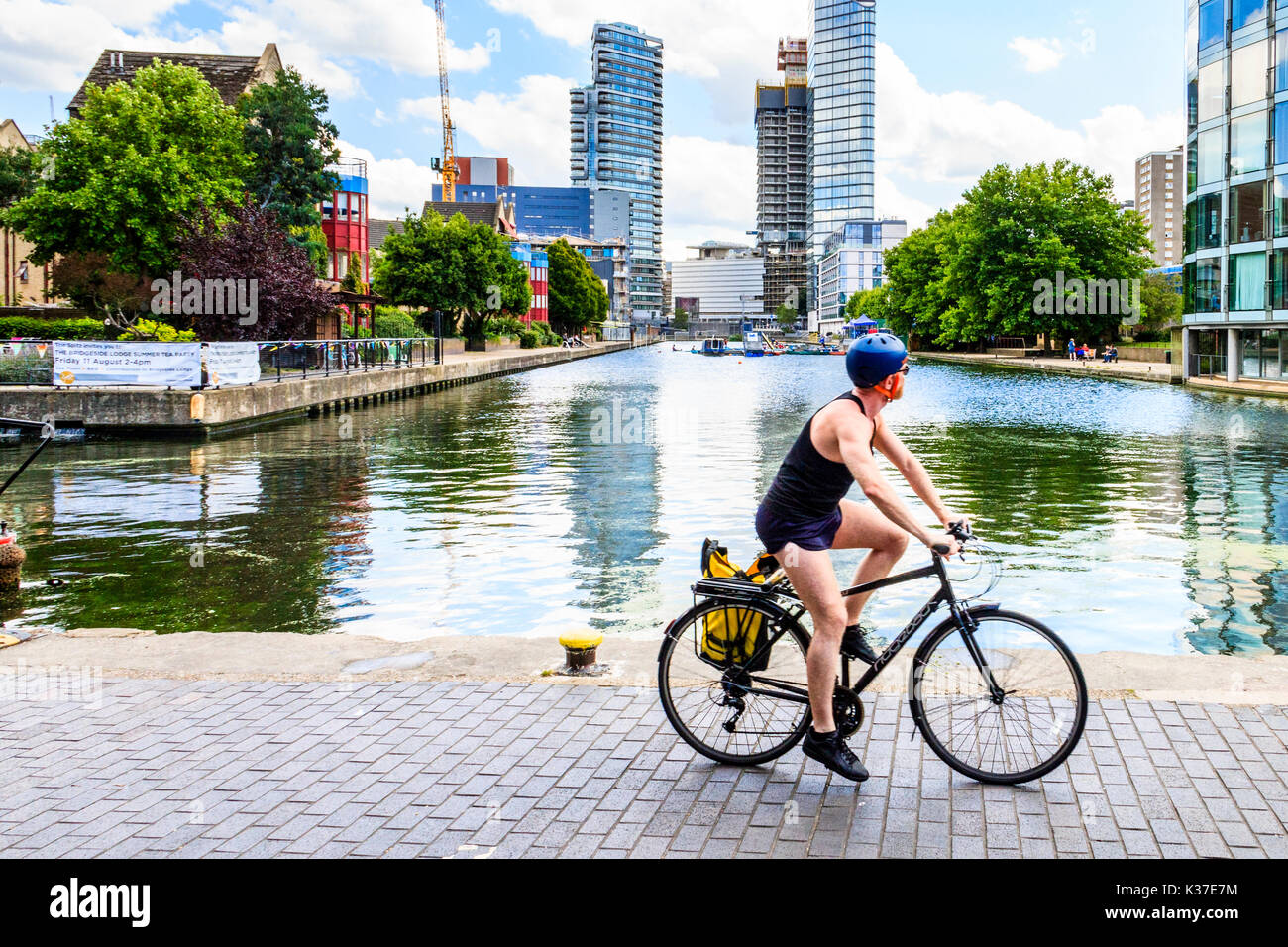 Un ciclista in shorts, gilet e casco che cavalcano una bicicletta sull'alzaia del canale di Regent a City Road Basin, Islington, Londra, UK, in una calda giornata estiva Foto Stock
