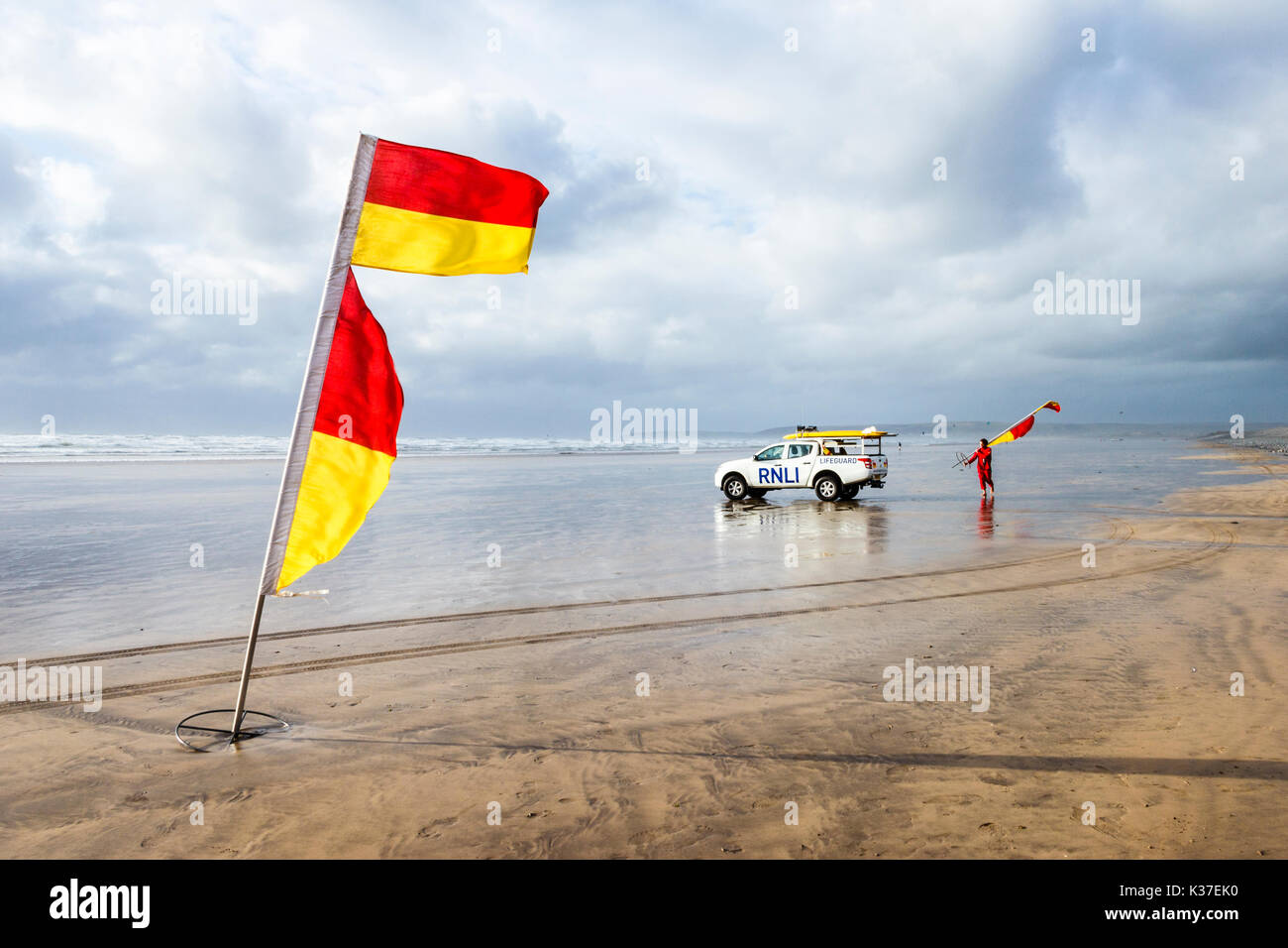 Rosso e giallo di avvertimento e bandiera RNLI bagnino di salvataggio con il veicolo sulla spiaggia sabbiosa di Condino Con la bassa marea su un nuvoloso e ventoso giorno di estate Foto Stock