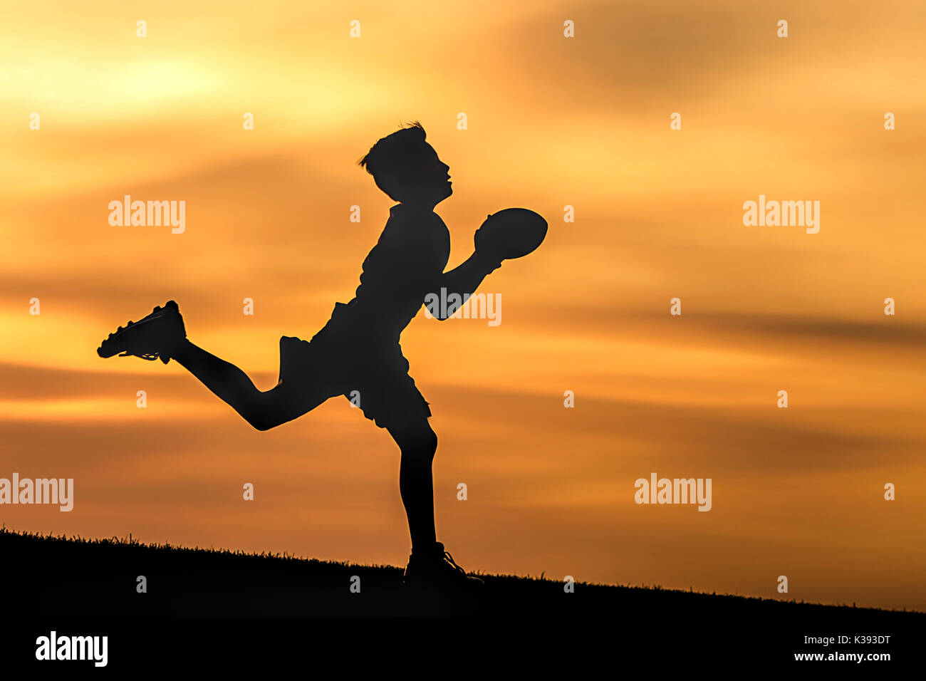 Un giovane ragazzo in silhouette è giocare a calcio al tramonto contro un cielo arancione. Foto Stock