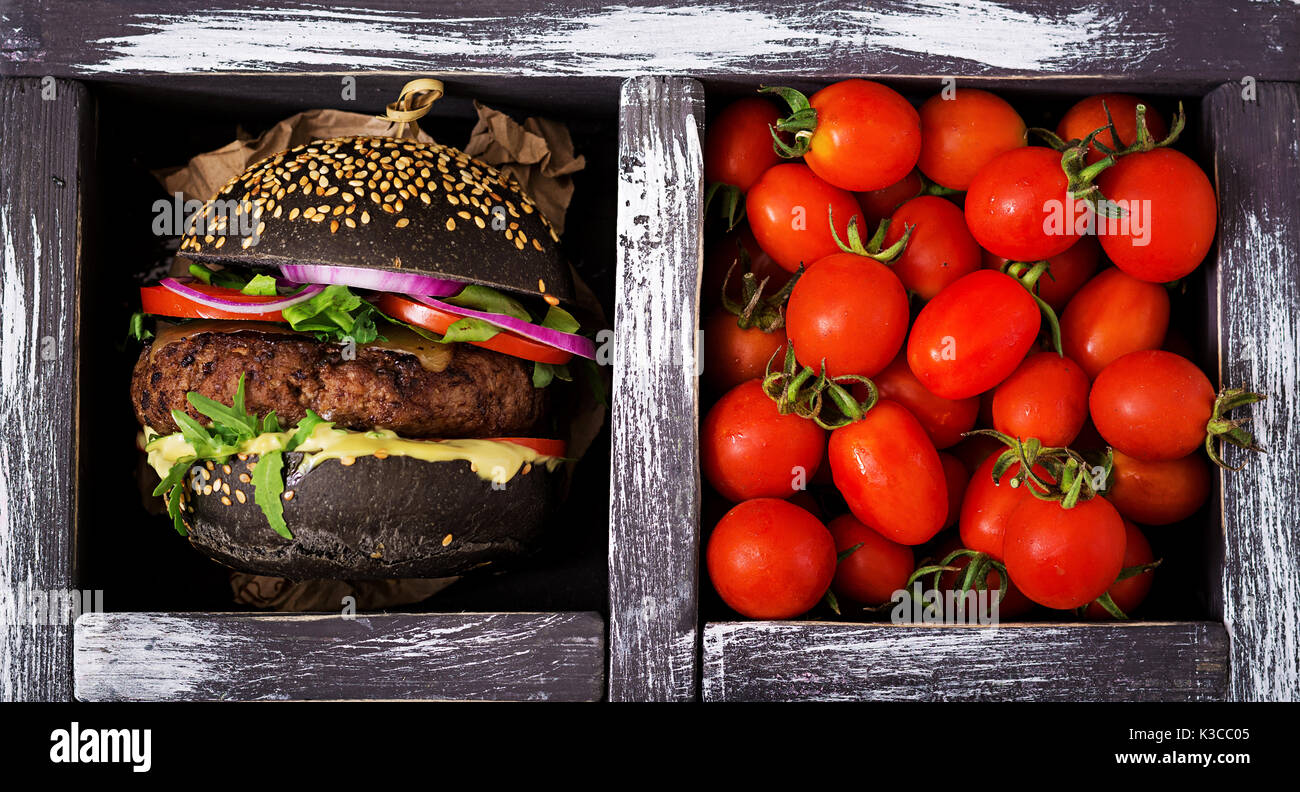 Nero grande panino - nero hamburger con succose carni bovine burger, Formaggio, pomodoro e cipolla rossa nella casella su sfondo nero. Lay piatto. Vista superiore Foto Stock