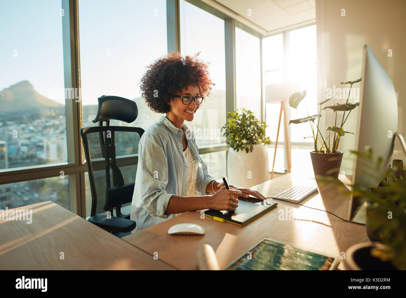 Felice femmina giovane designer che lavora con la tavoletta grafica e computer. Professionisti creativi a lavorare alla sua scrivania in ufficio. Foto Stock