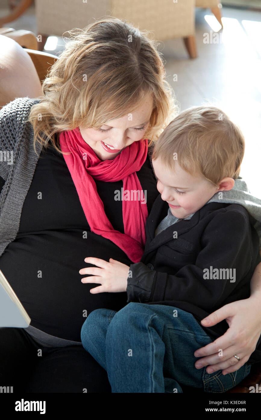 Sorridente madre incinta con il suo braccio intorno a suo figlio per fargli sentire la sua gravidanza pancia. Foto Stock