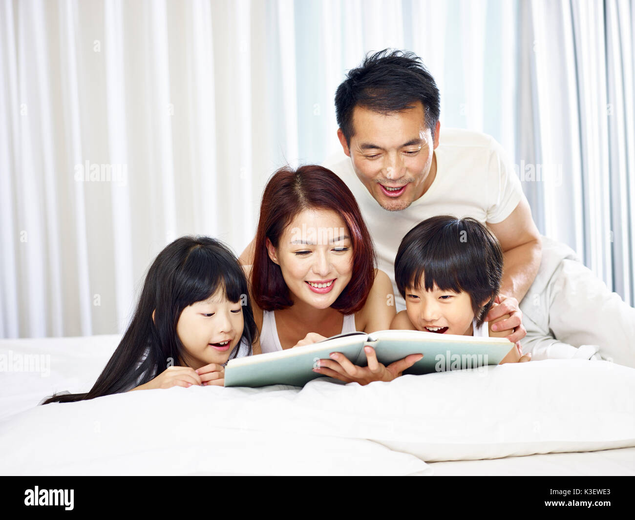 Felice famiglia asiatica con due bambini che giace sulla parte anteriore a letto a leggere un libro insieme. Foto Stock