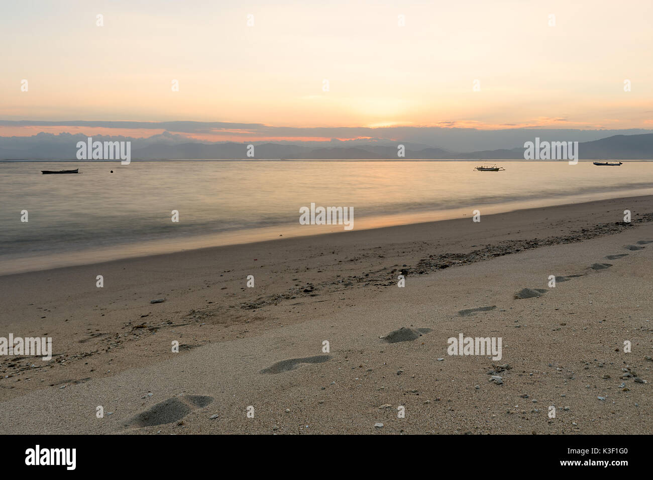 Piedi stampe nella sabbia dorata della spiaggia di Lakey, Sumbawa, Indonesia con 3 barche da pesca al tramonto. Foto Stock