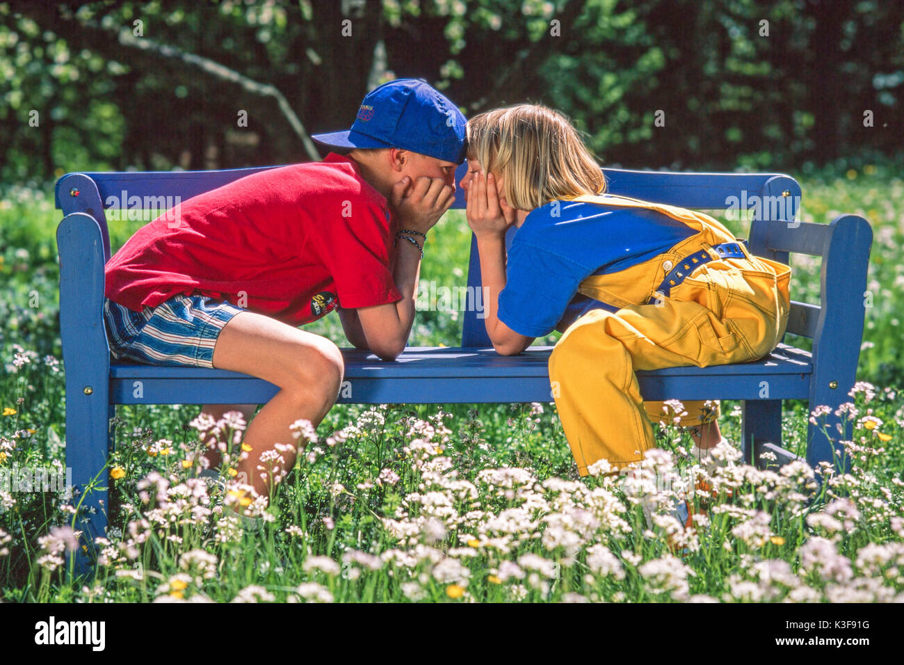 Un ragazzo e una ragazza sedersi in un giardino sul bracciolo di una panchina da giardino e whisper Foto Stock