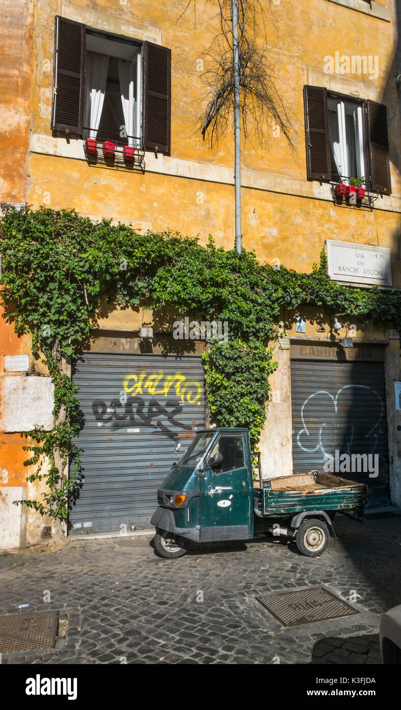 Vecchio verde scuro piaggio ape a tre ruote a veicolo commerciale parcheggiato di fronte a una facciata ricoperta nel centro storico della città di Roma Foto Stock
