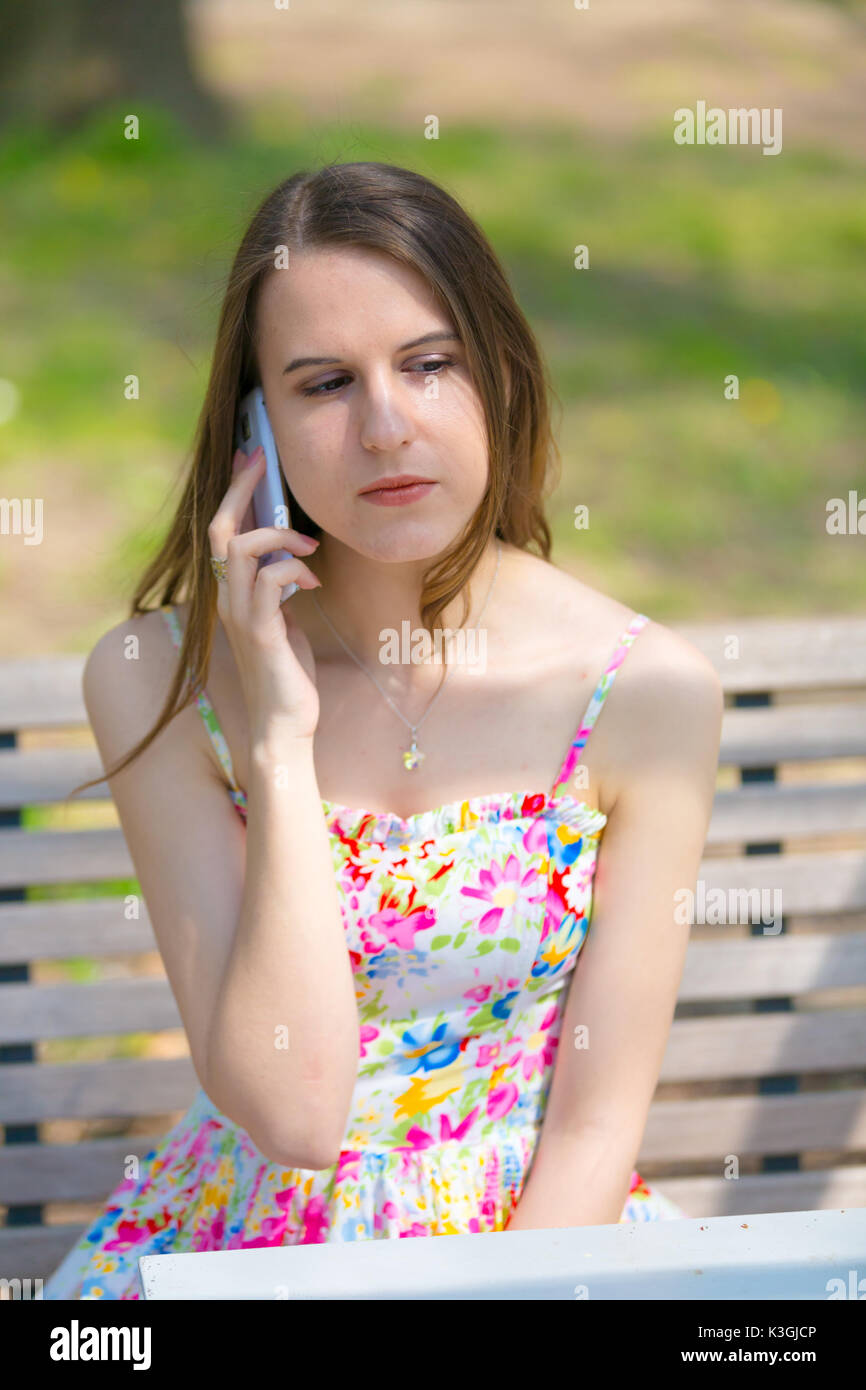 Ritratto di giovane bella donna con capelli lunghi in estate park, ragazza fiore di indossare abiti corti è utilizzando uno smartphone mentre è seduto su una panchina Foto Stock