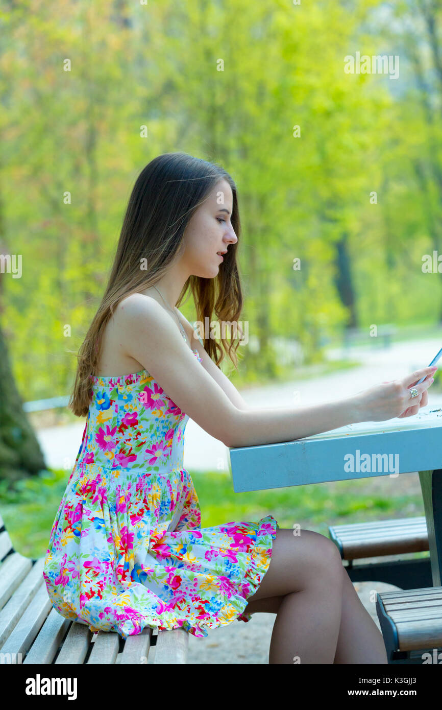Ritratto di giovane bella donna con capelli lunghi in estate park, ragazza fiore di indossare abiti corti è utilizzando uno smartphone mentre è seduto su una panchina Foto Stock