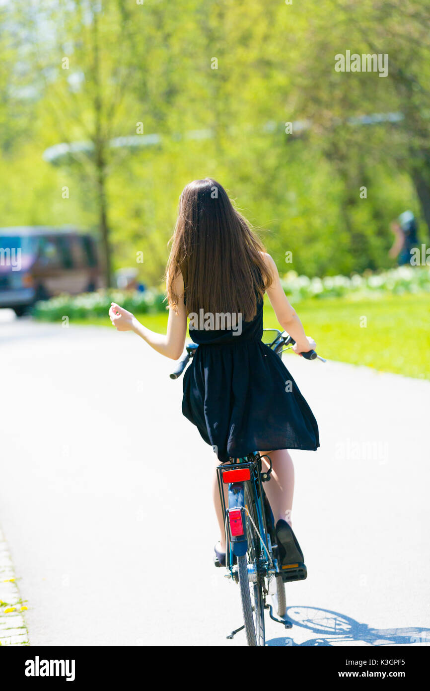 Vista posteriore di una ragazza in bicicletta che indossa sul nero abito corto. Giovane donna a cavallo lungo la strada a molla verde parco all'aperto. Sporty giovane ragazza a cavallo di un modulo di interconnessione BIC Foto Stock