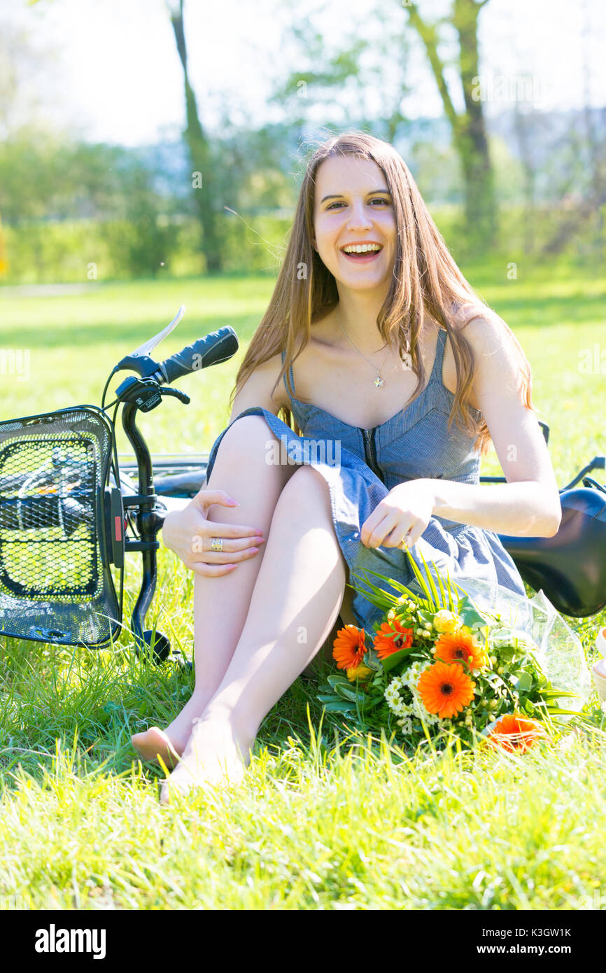 Giovane donna è seduta sul prato in un parco nei pressi di una bicicletta e un mazzo di fiori Foto Stock