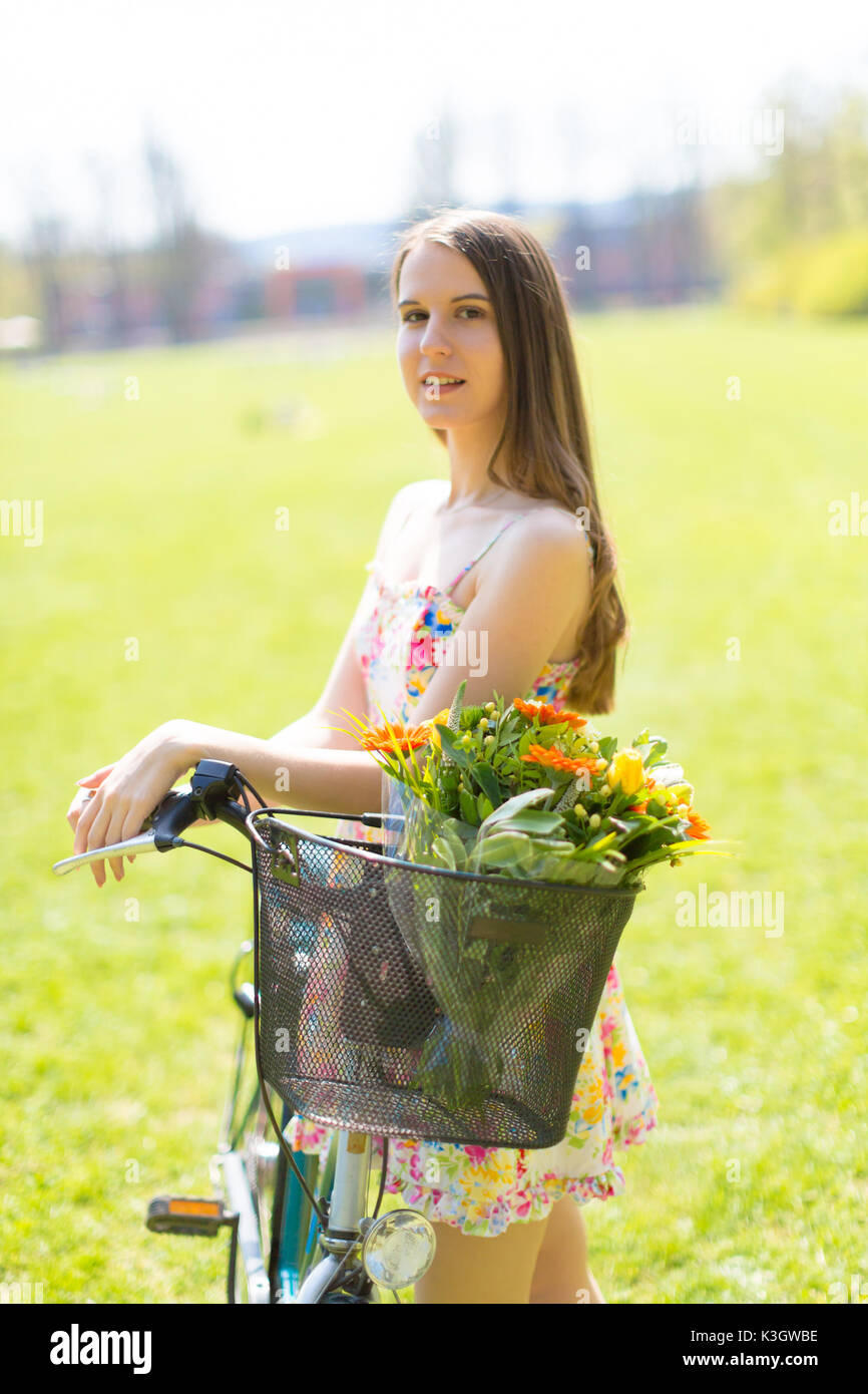 Ritratto di giovane bella donna con capelli lunghi in estate park. A fianco di una bicicletta con un cestino di fiori Foto Stock