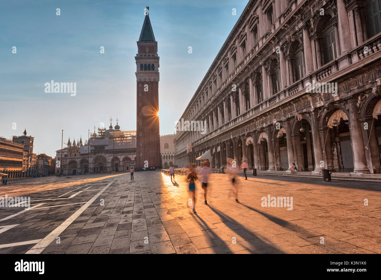 L'Europa, Italia, Veneto, Venezia. Le persone che eseguono in Piazza San Marco mentre il sole sorge da dietro il campanile San Marco Foto Stock