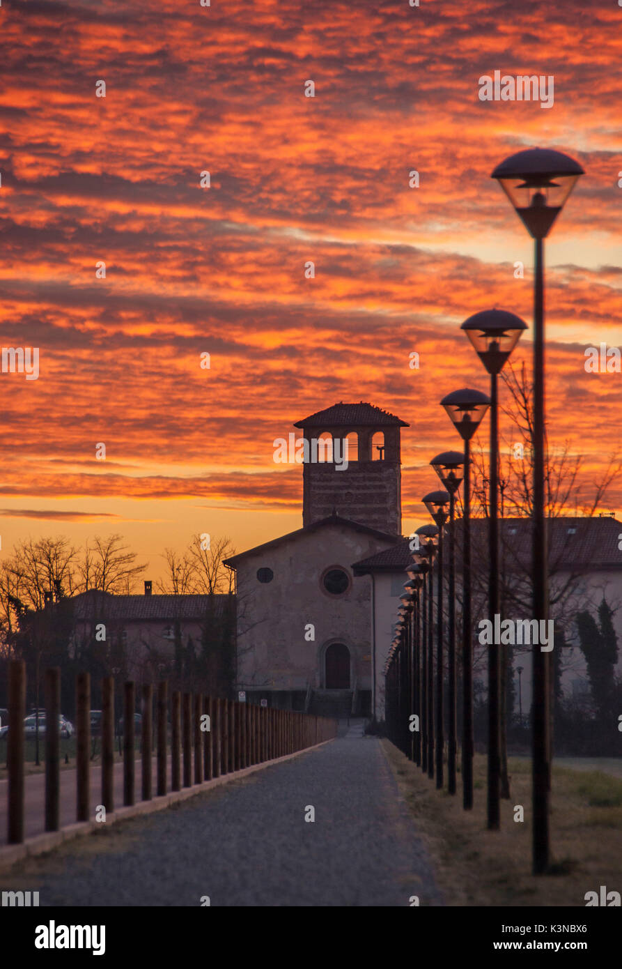 La strada, fiancheggiata da una fila di lampade stradali, che conduce alla chiesa di Santa Maria Maddalena nel borgo medievale di Cascina Camuzzago viste dell'alba con un cielo rosso e le nuvole. Bellusco, Monza e Brianza, Lombardia, Italia Foto Stock