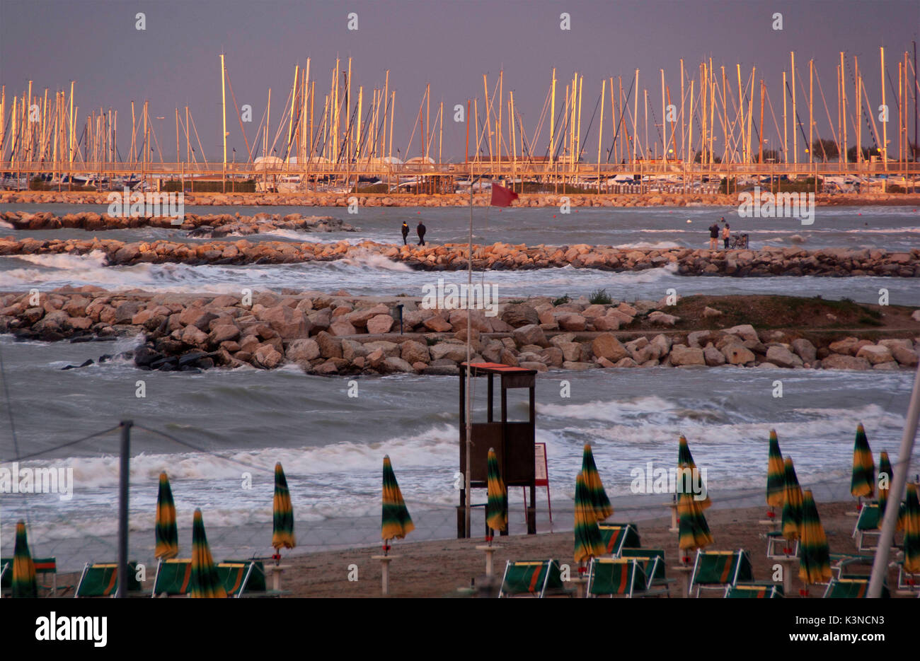 Il tramonto sulla spiaggia di Rimini illumina il mare mosso e il piccolo porto turistico con ormeggiate barche a vela sui bacini di pietre. Emilia Romagna, Italia Foto Stock