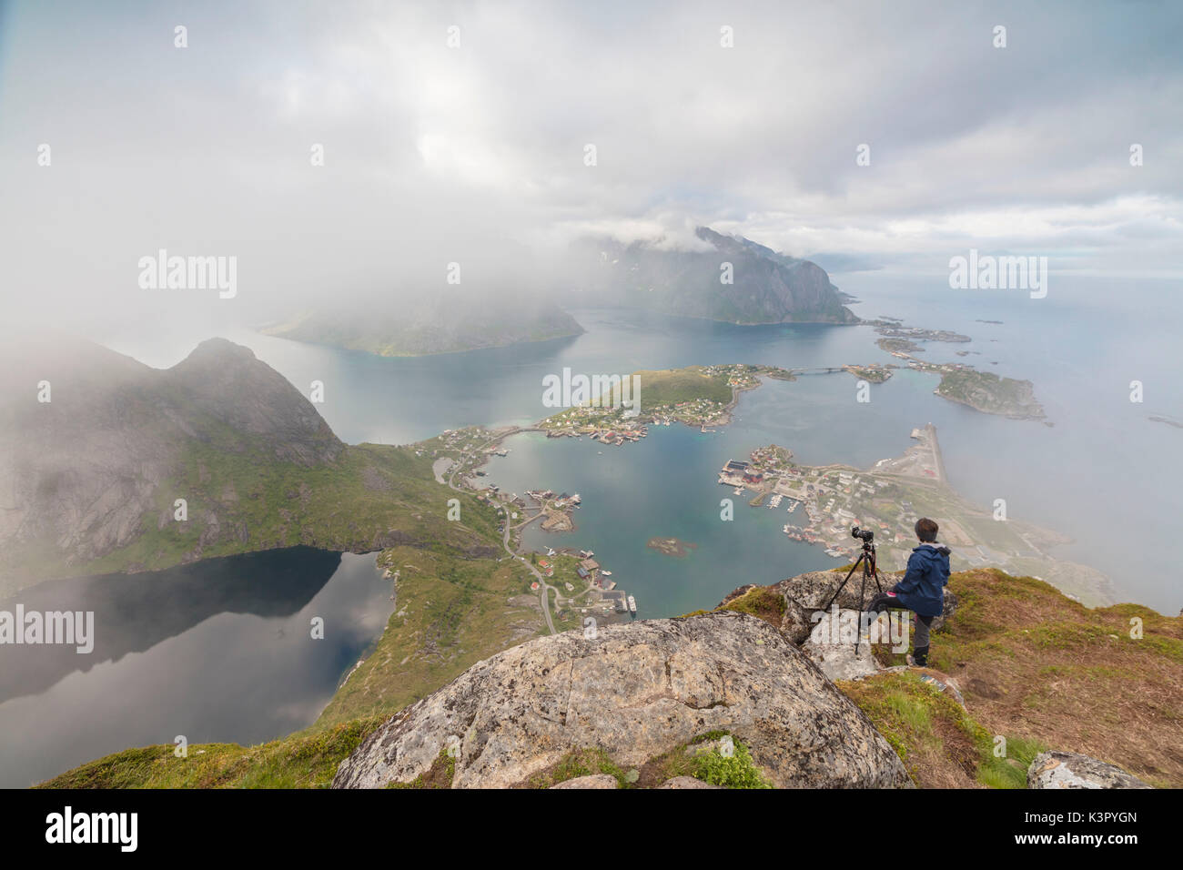 Fotografo sulla sommità del picco roccioso si ammira il blu del mare circondato da nebbia Moskenes Reinebringen Isole Lofoten in Norvegia Europa Foto Stock