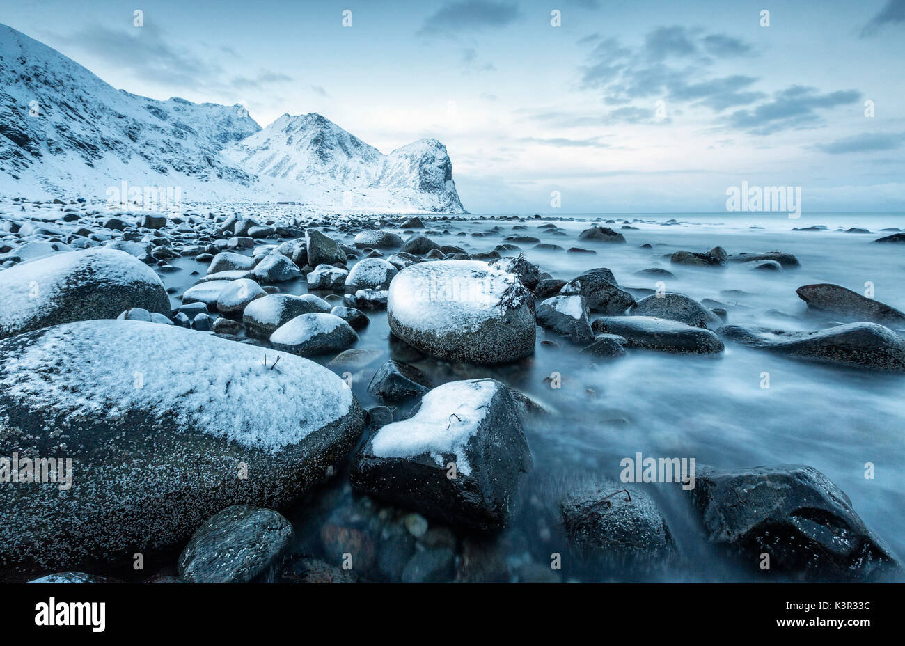 Rocce nel freddo del mare e delle montagne innevate sotto la luce blu del crepuscolo Unstad Isole Lofoten Norvegia del Nord Europa Foto Stock