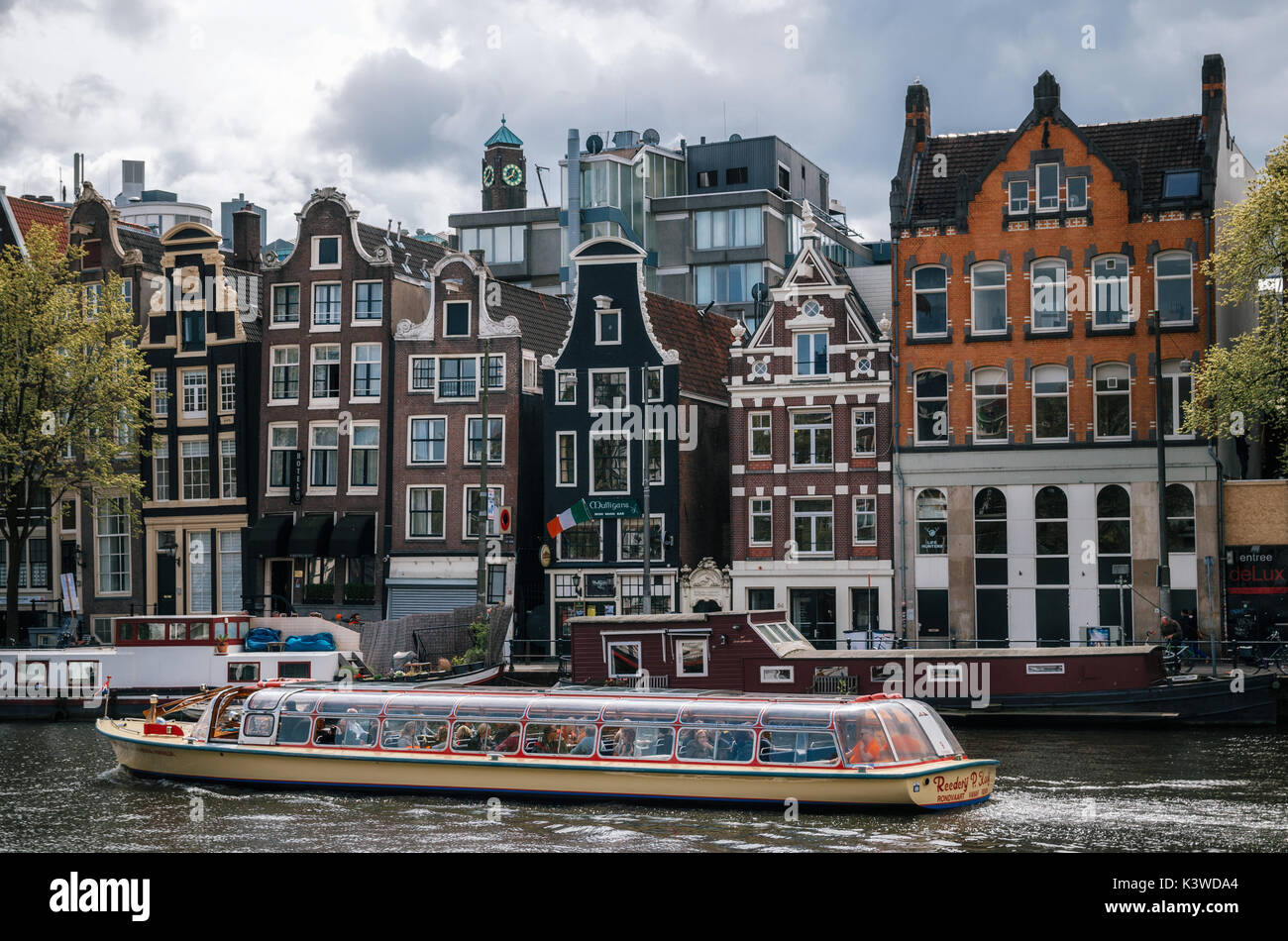 Amsterdam, Paesi Bassi - 27 April, 2017: Crociera imbarcazione turistica contro di case galleggianti, chiatte vivente e curve tipiche case olandesi con grandi finestre in B Foto Stock