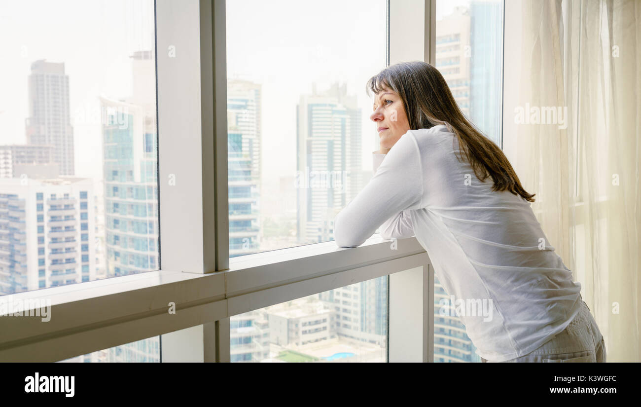 Donna matura è guardando fuori della finestra nel suo hotel/appartamento Foto Stock