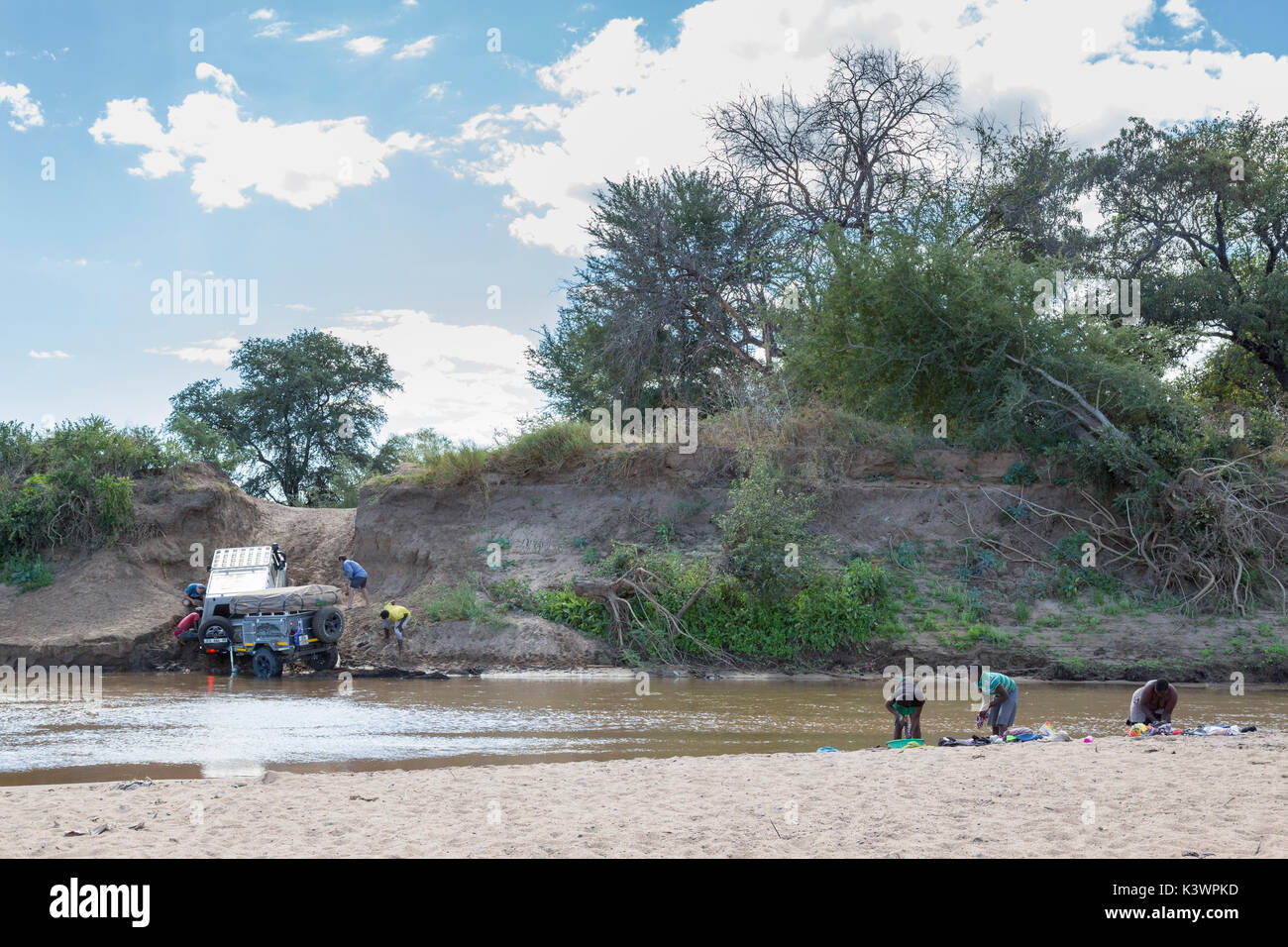 Landcrusier con rimorchio lottando per attraversare il fiume Limpopo durante la salita di una sponda ripida con tre donne locali facendo loro lavaggio cercando su Foto Stock