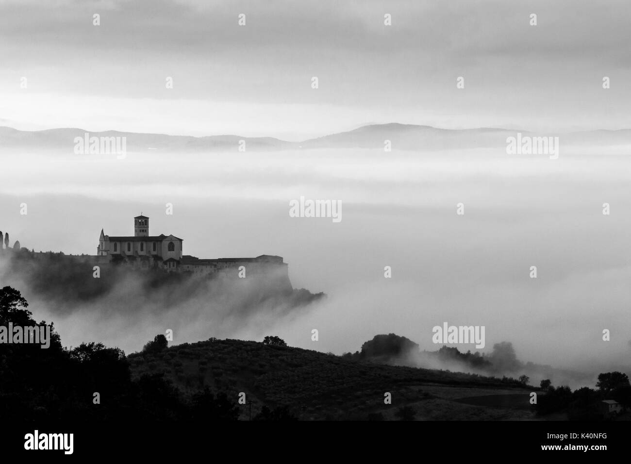 Bella vista monocromatica di San Francesco in Assisi (Umbria, Italia), sopra un mare di nebbia all'alba, con le colline e gli alberi in primo piano Foto Stock