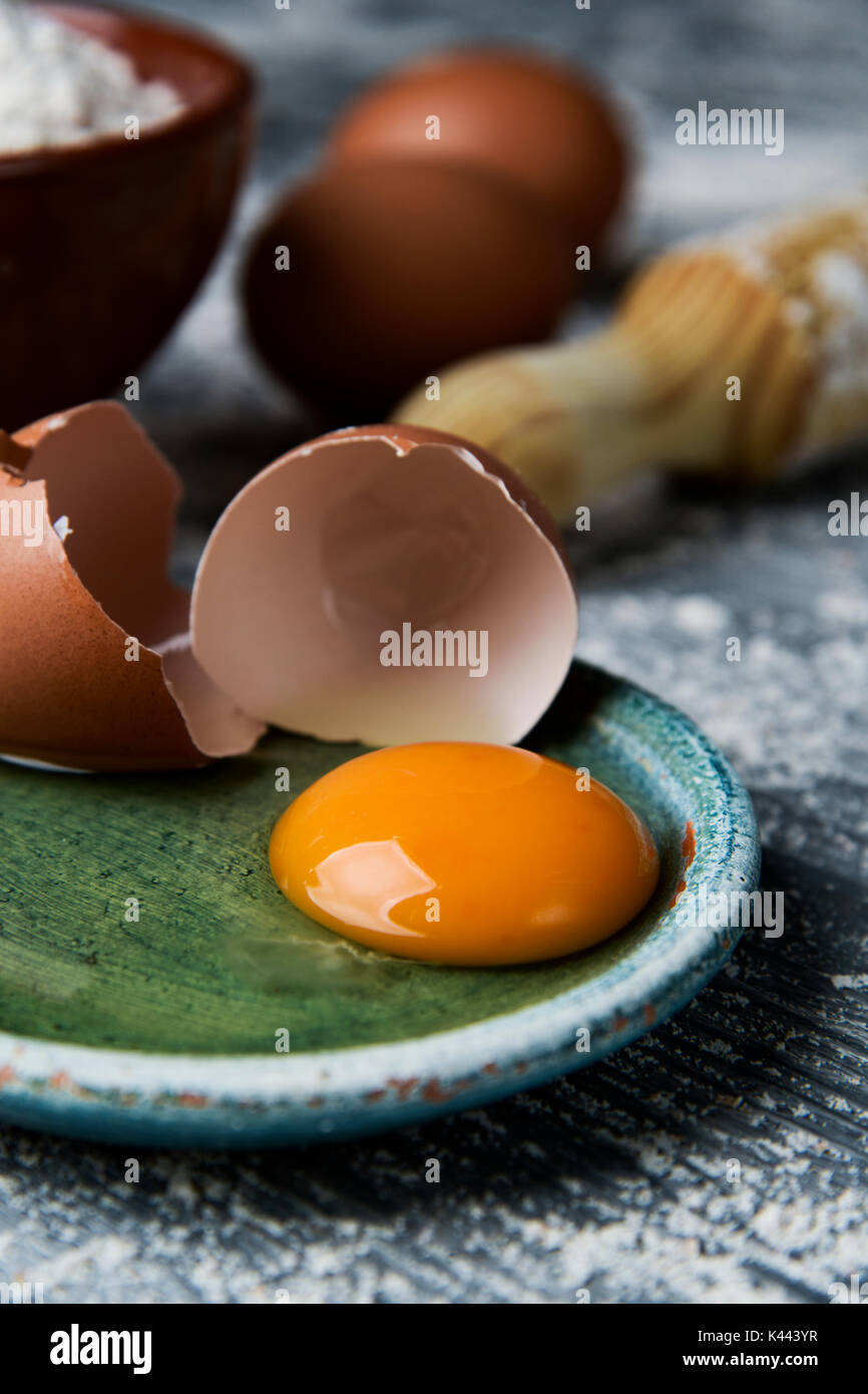 Primo piano di un uovo rotto in una piastra verde, un vaso di terracotta con farina, alcune uova intere e una laminazione di legno pin su un tavolo rustico cosparso con f Foto Stock