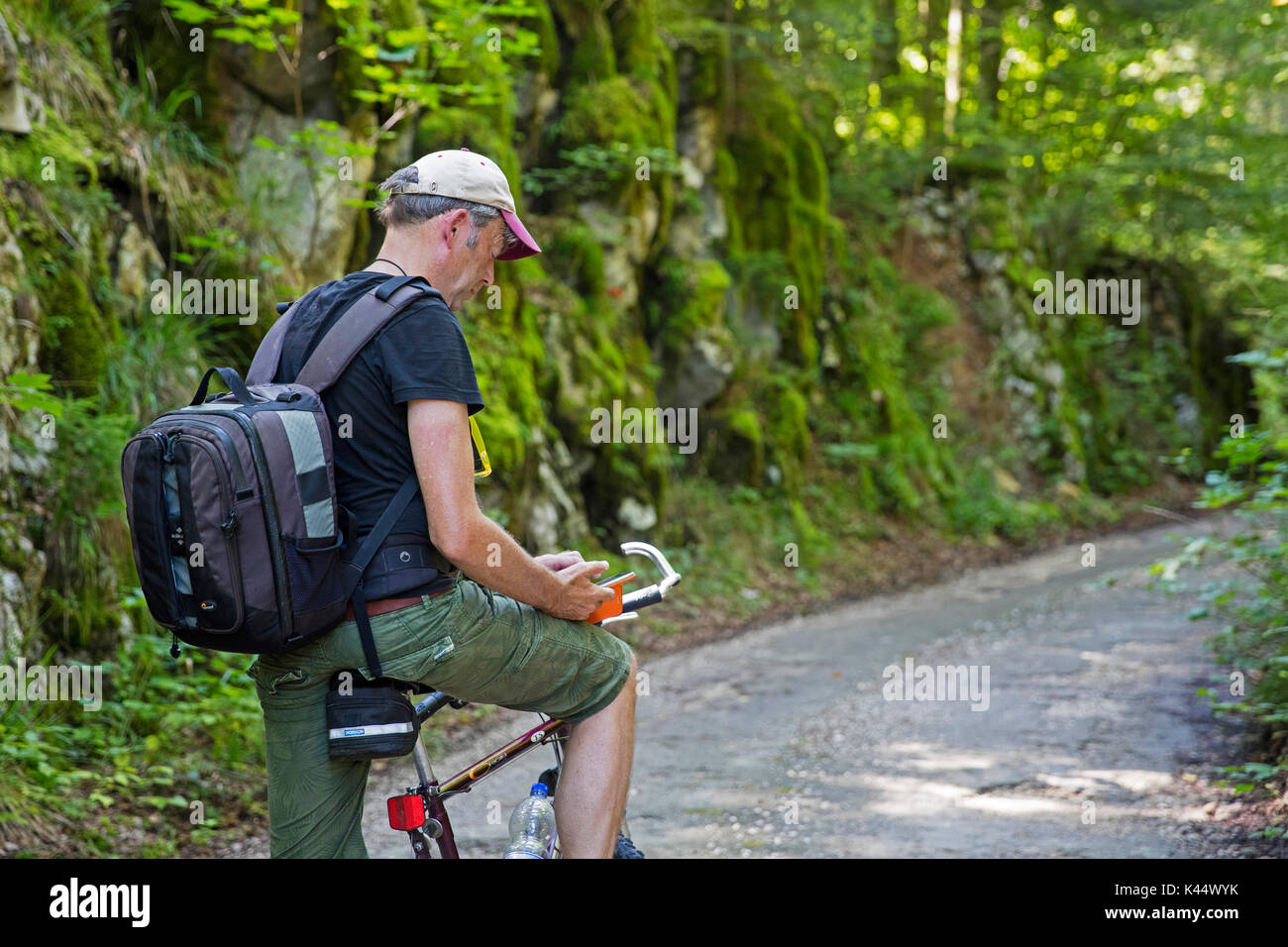Ciclista con zaino in sella ad una bicicletta fermarsi a prendere una pausa per sms e smartphone di lettura lungo una strada forestale in estate Foto Stock