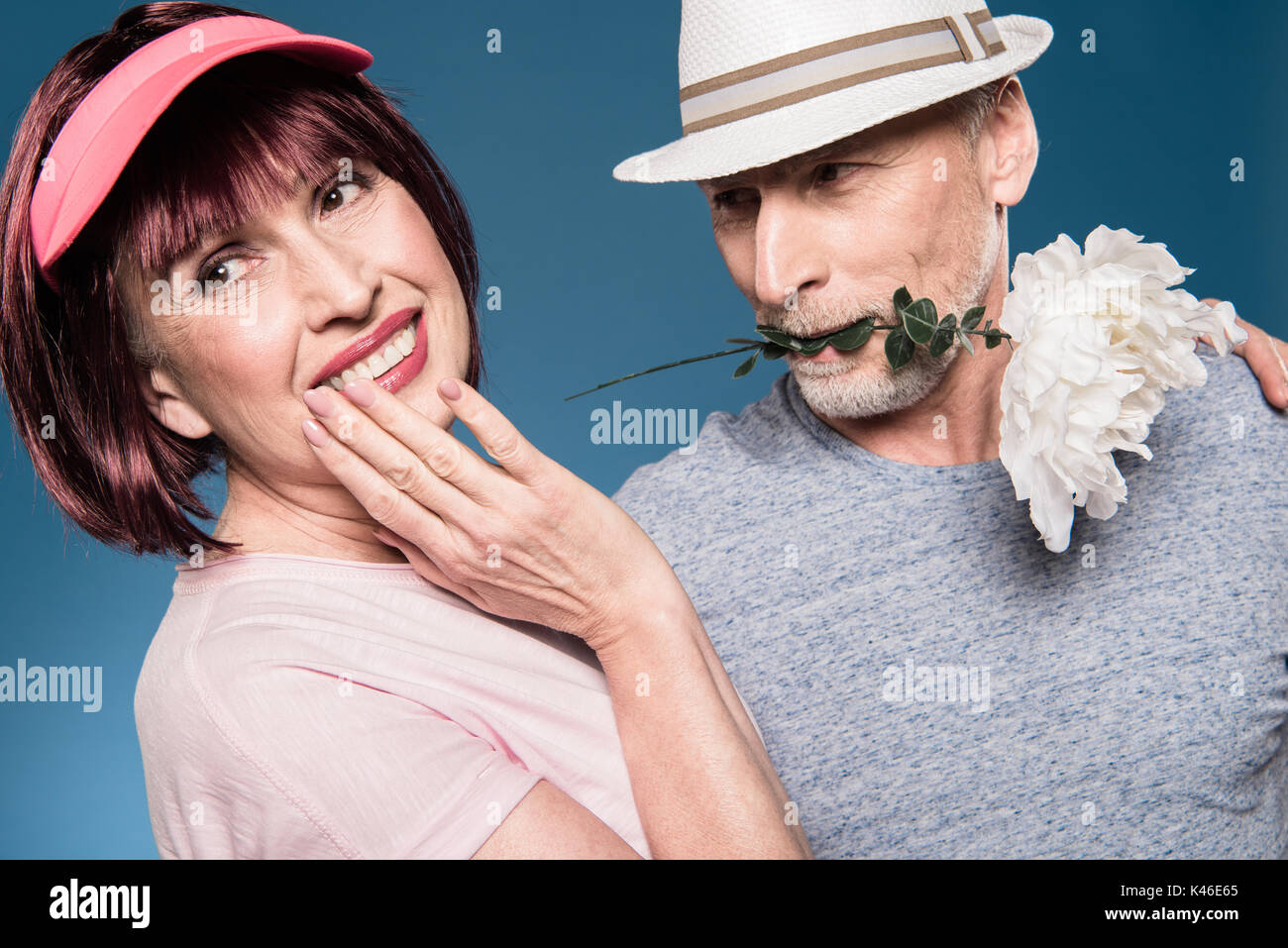 Elegante coppia di anziani che abbraccia e posa con fiore bianco isolato su blu Foto Stock