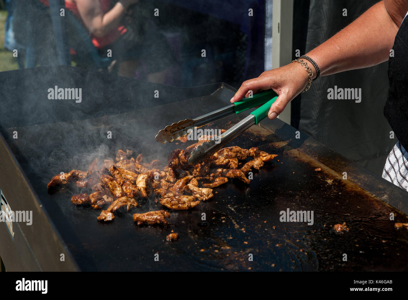 Pollo alla griglia su piastra calda cucinando in primo piano con le mani dello chef e le pinze che sfrigolano la carne Foto Stock