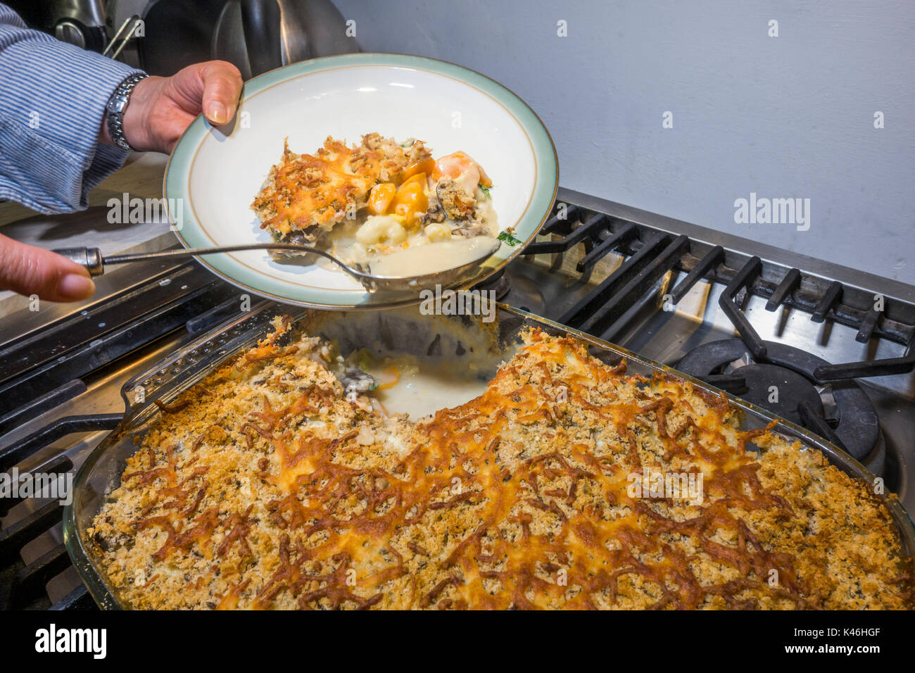 Donna con le mani in mano il trasferimento a caldo cuocere verdure, con mollica di pane e formaggio croccante topping, da un vetro piatto su una piastra utilizzando un grande cucchiaio. Foto Stock