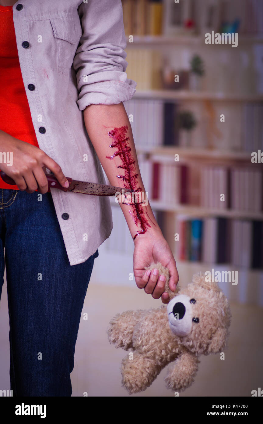 In prossimità di una giovane donna depressiva con il suo braccio lo spurgo, tenendo in mano un orsacchiotto di peluche e un coltello nella sua altra, in uno sfondo sfocato Foto Stock