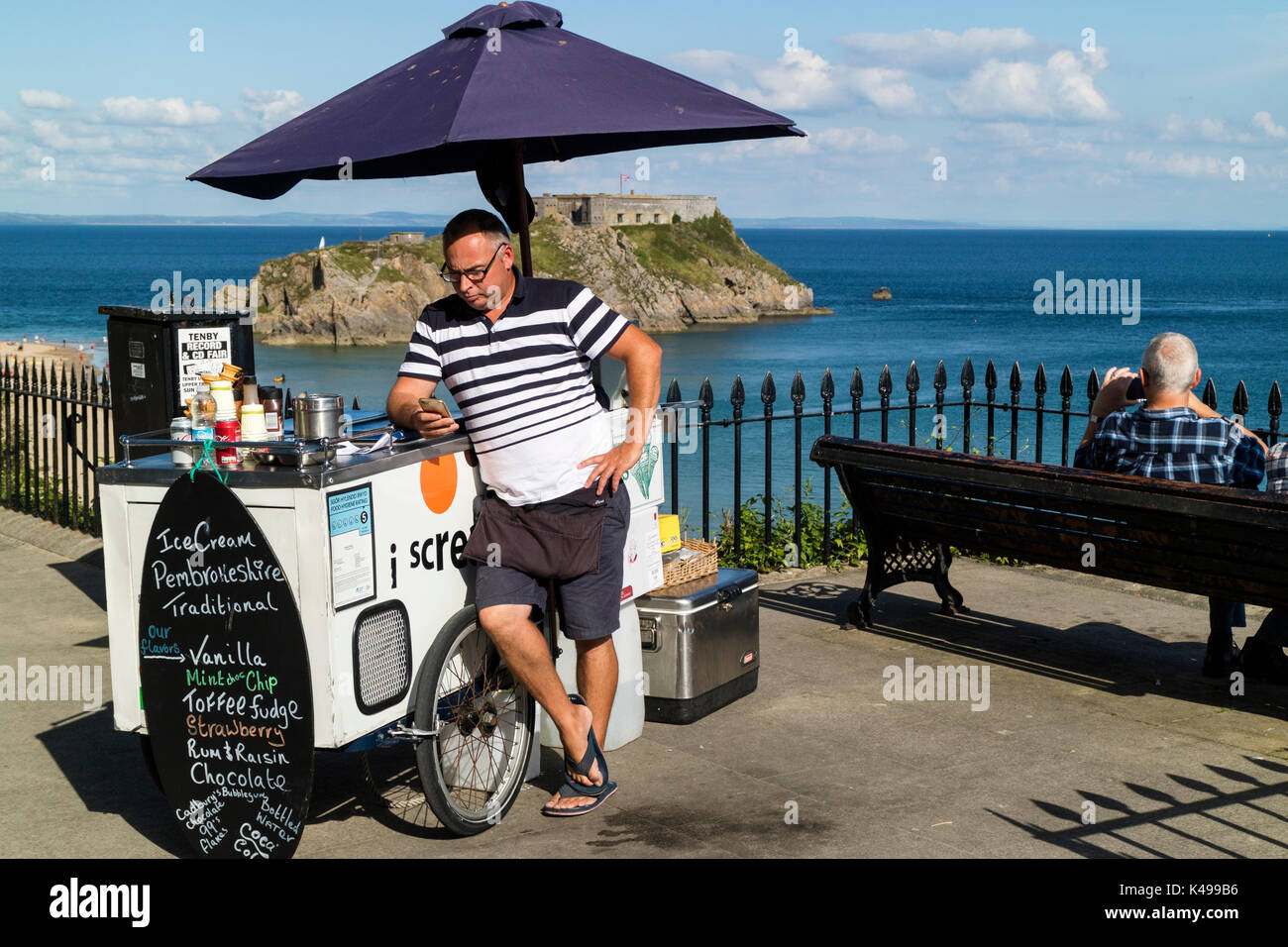 Un uomo di vendita di gelato da una bicicletta sul fronte mare in Tenby, Galles controlla il suo mobile / Telefono cellulare con Santa Caterina di isola in background Foto Stock