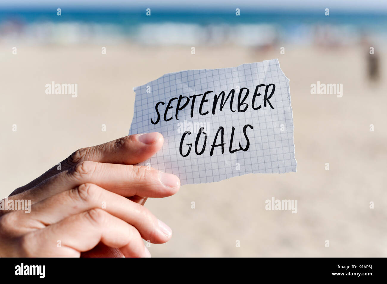 Primo piano della mano di un giovane uomo caucasico tenendo una nota con il testo obiettivi settembre scritto in esso, di fronte alla spiaggia Foto Stock