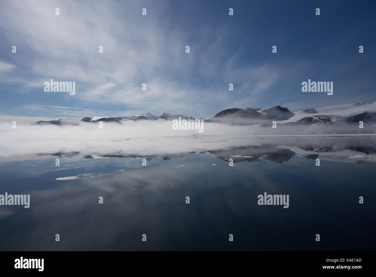 La costa settentrionale di Spitsbergen, Svalbard si riflette nelle acque calme dell'Oceano Artico, con una fascia di nebbia che si forma sul mare. Foto Stock