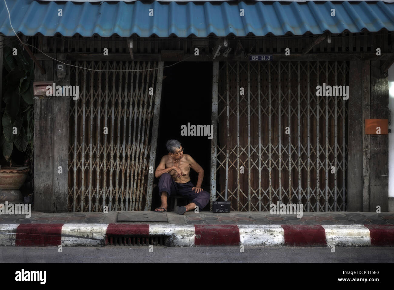 Thailandia casa urbana. Anziano tailandese uomo seduto sul marciapiede presso la sua casa tradizionale, con pesanti cassettini. Thailandia Sud-est asiatico Foto Stock