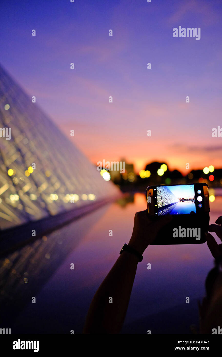 Un turista di scattare una foto sul loro smartphone del Louvre di Parigi di notte come il sole tramonta Foto Stock