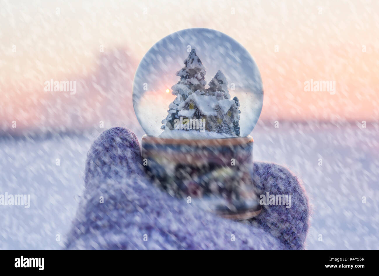 La ragazza di guanti a maglia tenendo palla di vetro con firtrees, casa e la neve artificiale con paesaggio invernale con la caduta di neve in background Foto Stock