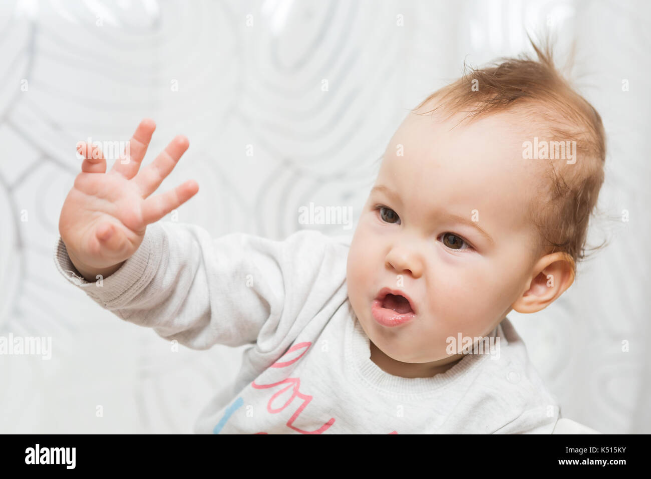 Undici mesi baby ragazza seduta in uno sgabello facendo facce buffe nella fotocamera e sollevando il suo braccio destro in aria come se si tenta di interrompere qualcuno o Foto Stock