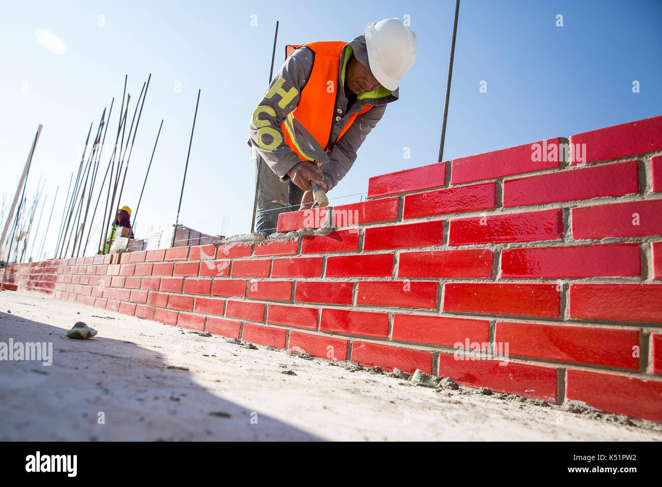 Operaio edile concentrato che costruisce un muro di mattoni rossi e Malta in un cantiere all'aperto, indossando l'attrezzatura di sicurezza e riflettendo il duro lavoro e il duro lavoro Foto Stock