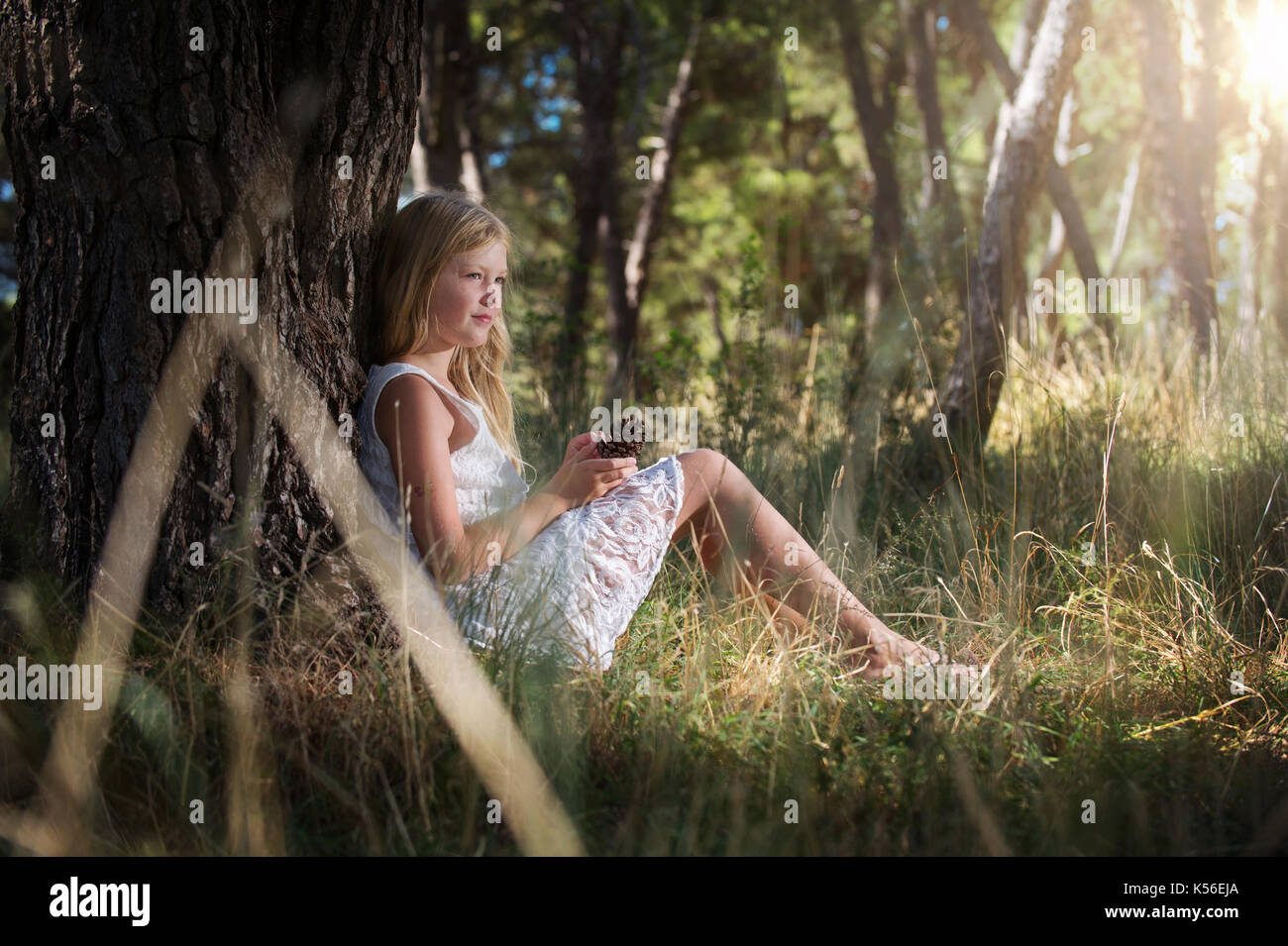 Lunghi capelli biondi carina ragazza in abito bianco, seduto appoggiato la schiena contro albero con cono di pino nelle mani di fata si assomigliano. Foto Stock